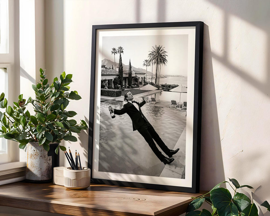 Framed black and white photograph of a person by a pool with palm trees, on a wooden shelf with plants.
