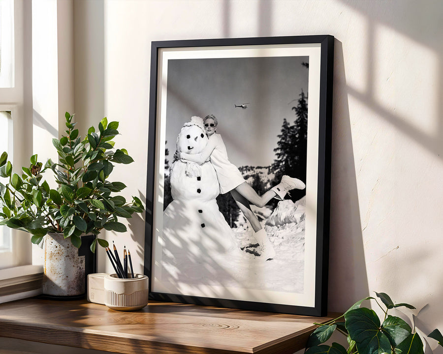 Framed black and white photo of a person with a dog snowman on a shelf with plants.