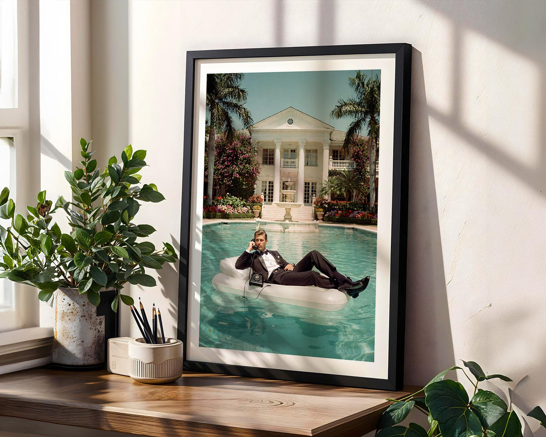 Framed artwork of a man on an inflatable pool float by a pool, leaning against a wall with plants.