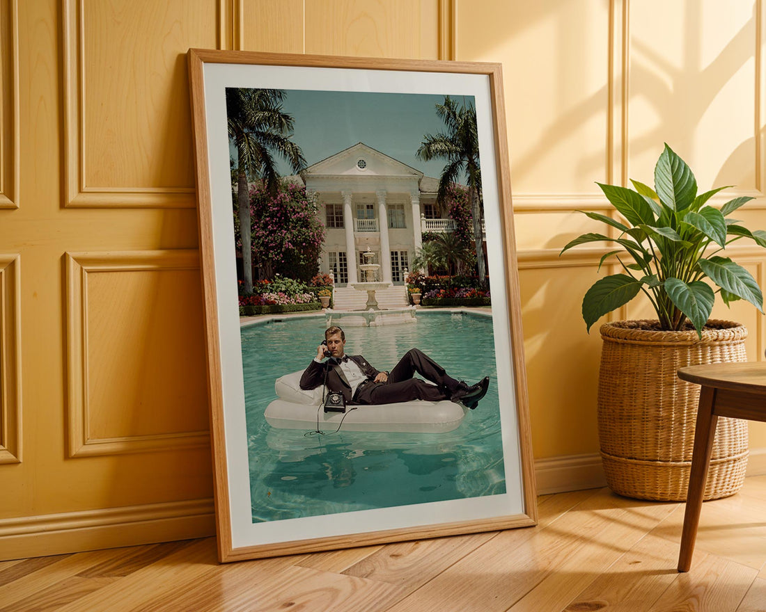 Framed photograph of a man on an inflatable pool float by a pool, leaning against a wooden wall with a plant and table.