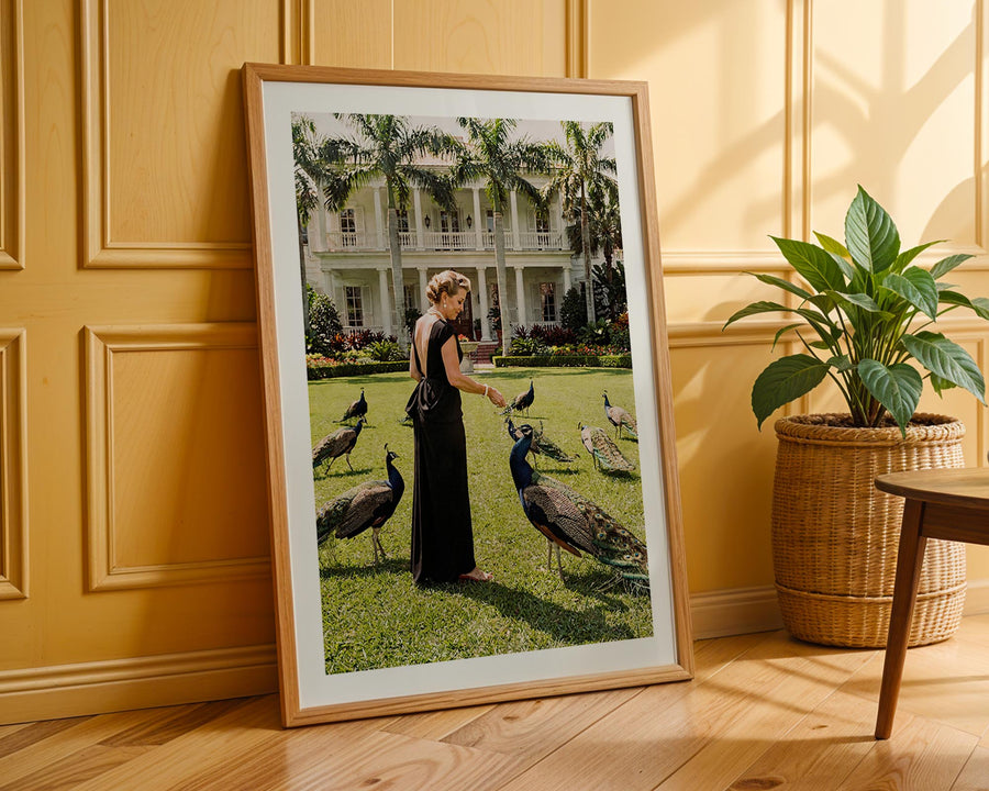 Framed photograph of a woman with peacocks in a garden setting, leaning against a wooden wall.