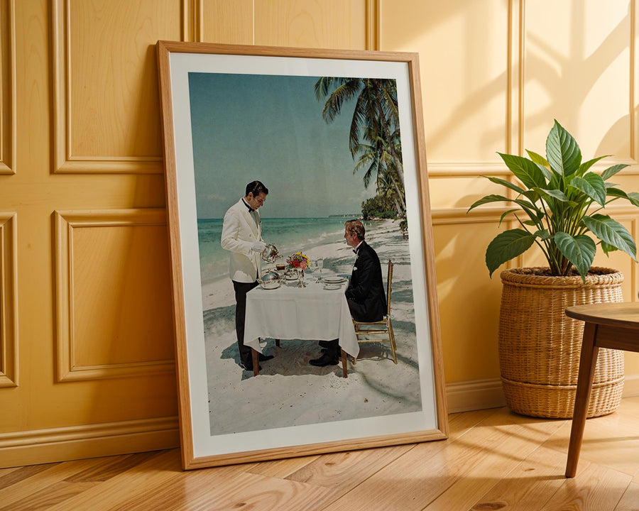 Framed photograph of two people at a beachside table with a plant and wooden floor in the background