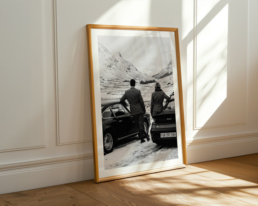 Framed black and white photograph of two people by a car on a mountain road, placed on a wooden floor with sunlight casting shadows.