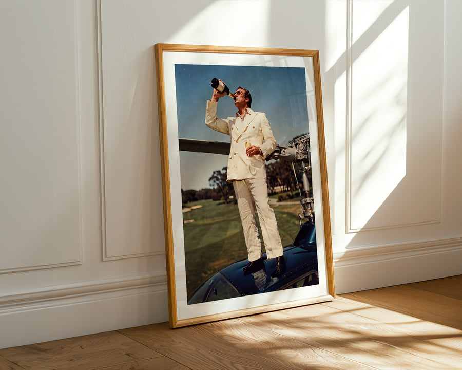 A framed art print leaning against a white paneled wall on a light wood floor, showing a vintage photograph of a man in a cream suit standing on a blue car on a golf course.