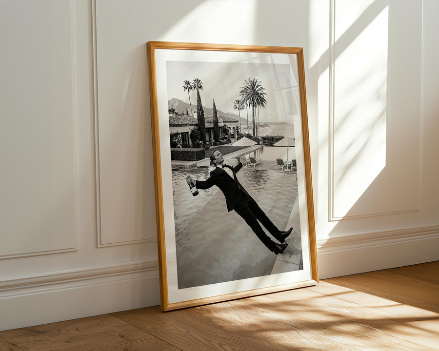 Framed black and white photograph of a person in a pool with palm trees and a building in the background, placed on a wooden floor.
