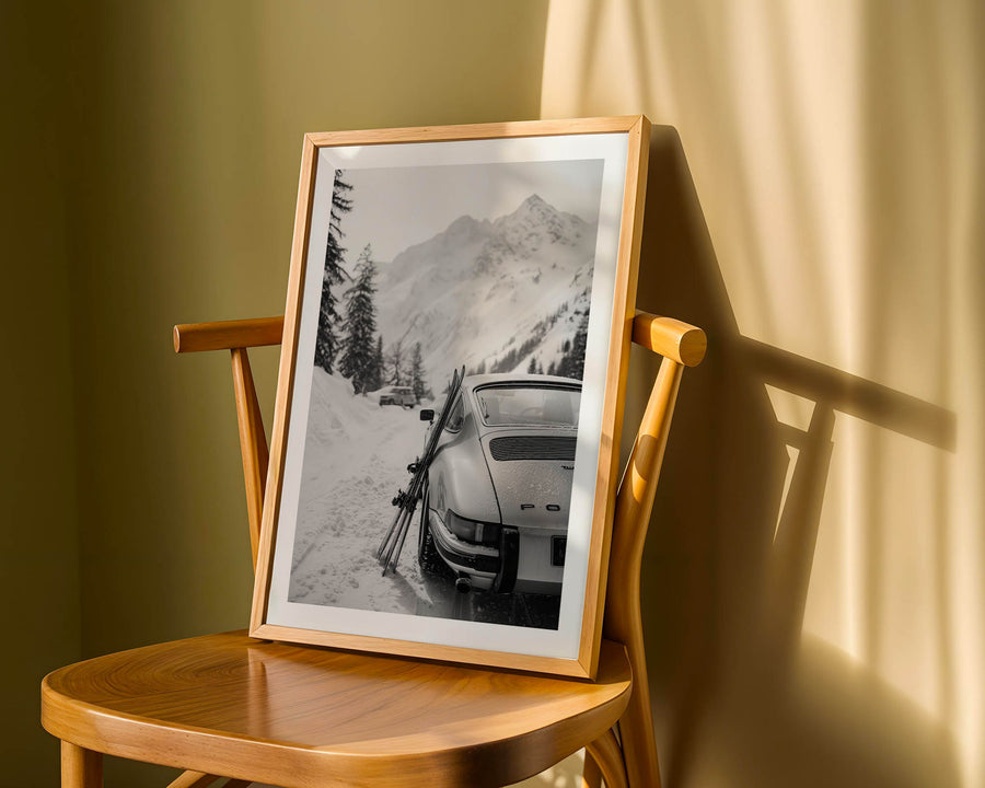 Framed photograph of a mountain landscape with a Porsche 911 on a wooden chair against a yellow wall.