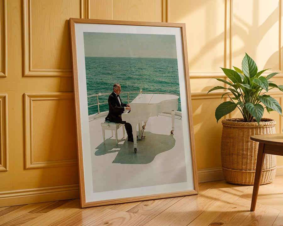 Framed photograph of a person playing a piano on a boat, leaning against a wooden wall with a plant beside it.