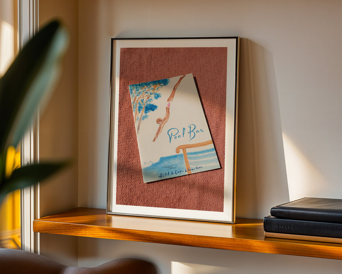 Framed artwork on a shelf with a plant and books in the background. Hôtel du Cap-Eden-Roc