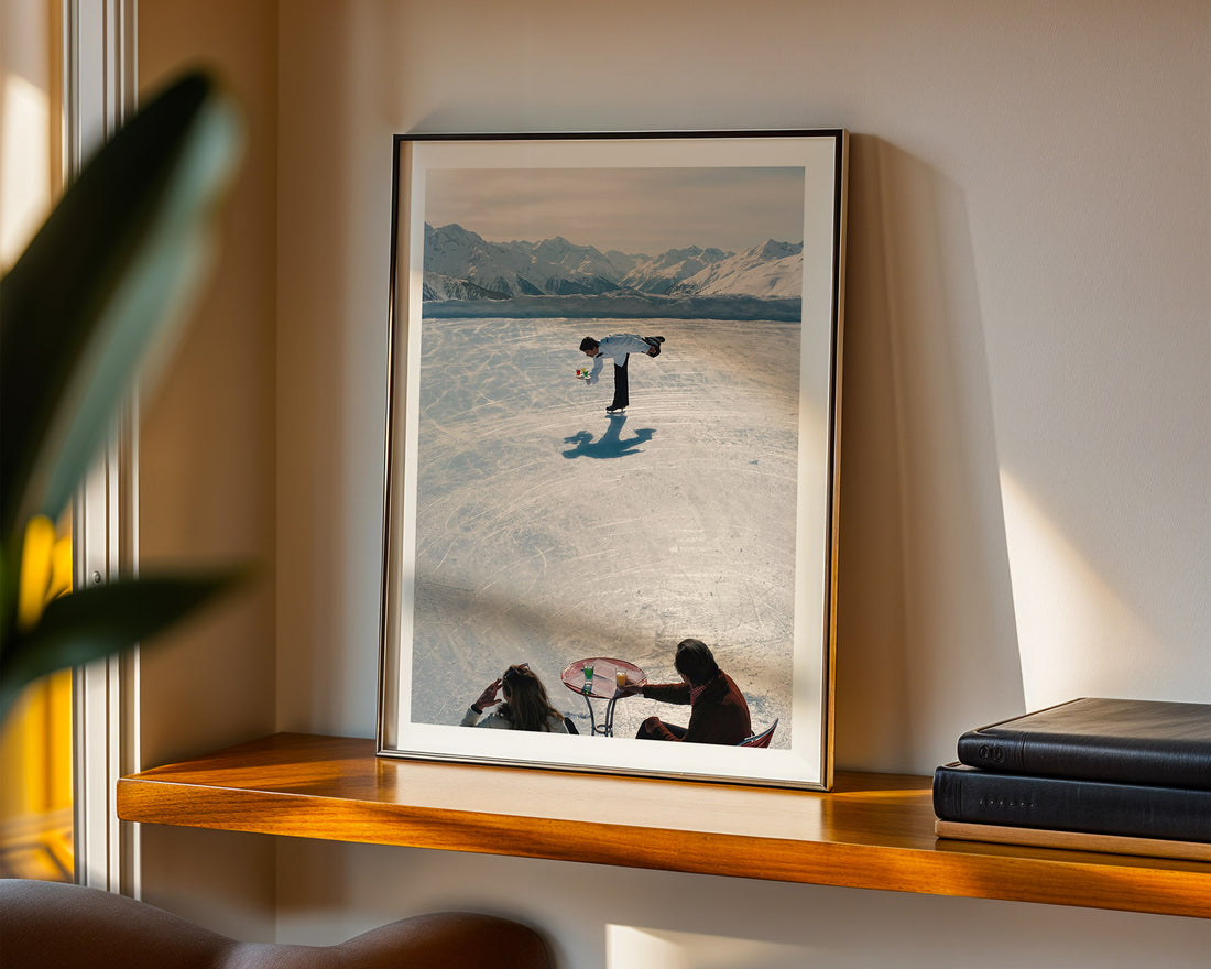 Framed artwork of a person ice-skating in Gstaad with mountains in the background, displayed on a wooden shelf.