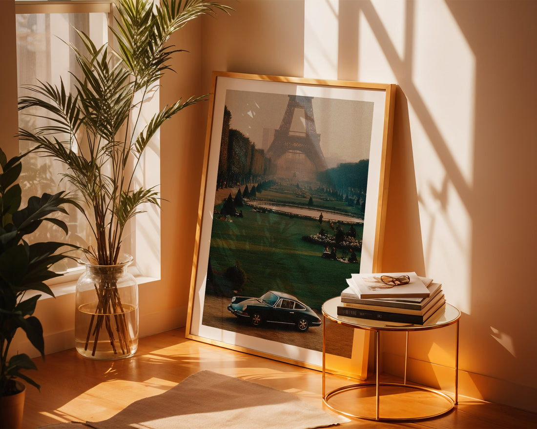 Framed artwork of the Eiffel Tower and Parisian scene on a wall with a plant and small table in the foreground.