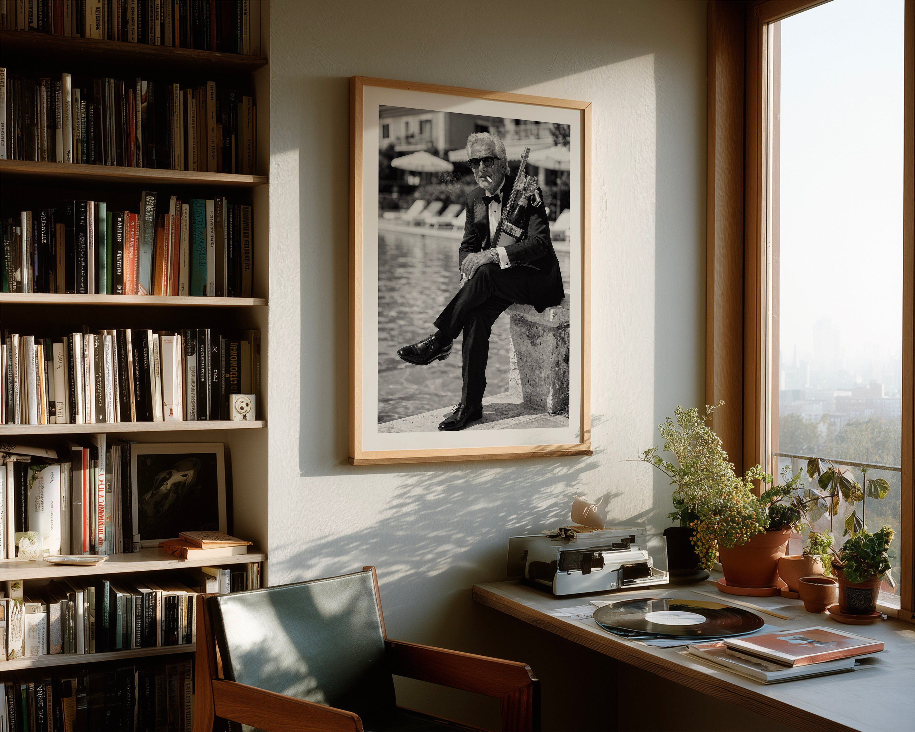 A framed black and white art print of a man sitting on a stone by a river, displayed on a wall in a warm, sunlit study with a bookshelf, desk, and plants.