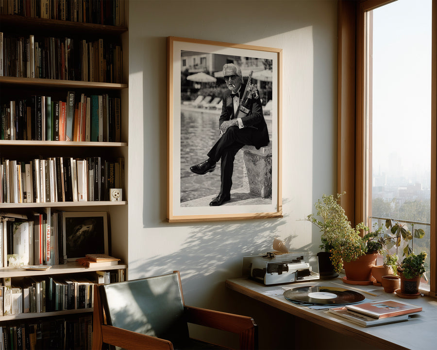 A framed black and white art print of a man sitting on a stone by a river, displayed on a wall in a warm, sunlit study with a bookshelf, desk, and plants.