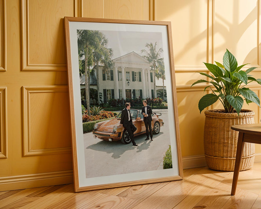 Framed photograph of two men by a vintage car on a wooden floor with a plant in the background.