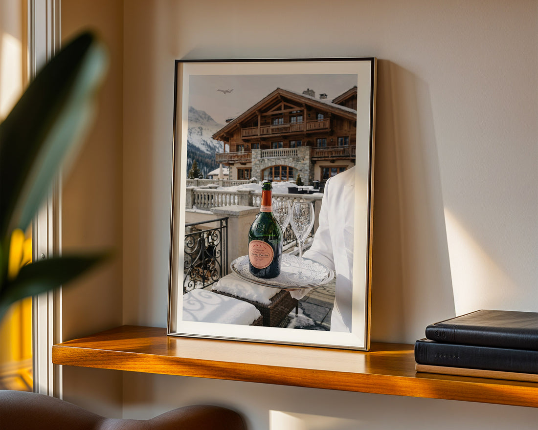 Framed artwork of a mountain lodge  scene in Gstaad with a bottle of champagne on a ledge.