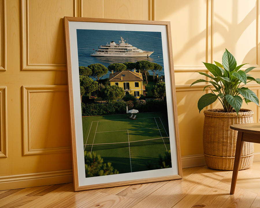 Framed artwork of a yacht and house on a tennis court against a yellow wall.
