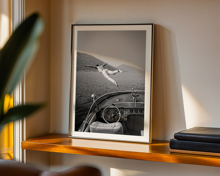Framed black and white photograph of a person lying on a car in a desert setting, displayed on a wooden shelf.