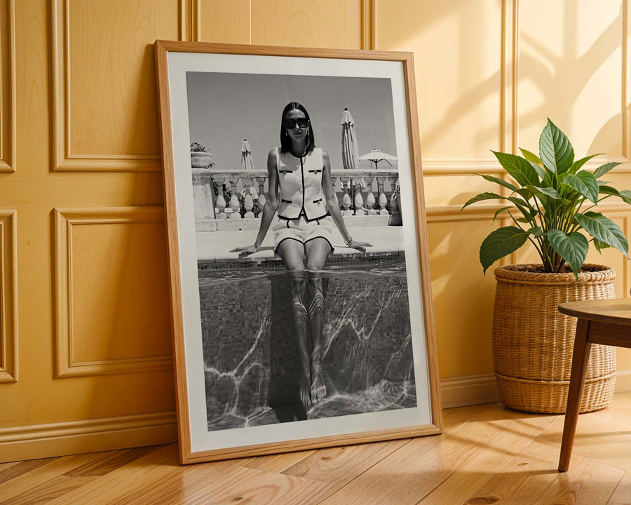 Framed black and white photograph of a woman sitting by a pool on a wooden floor with a plant in the background.