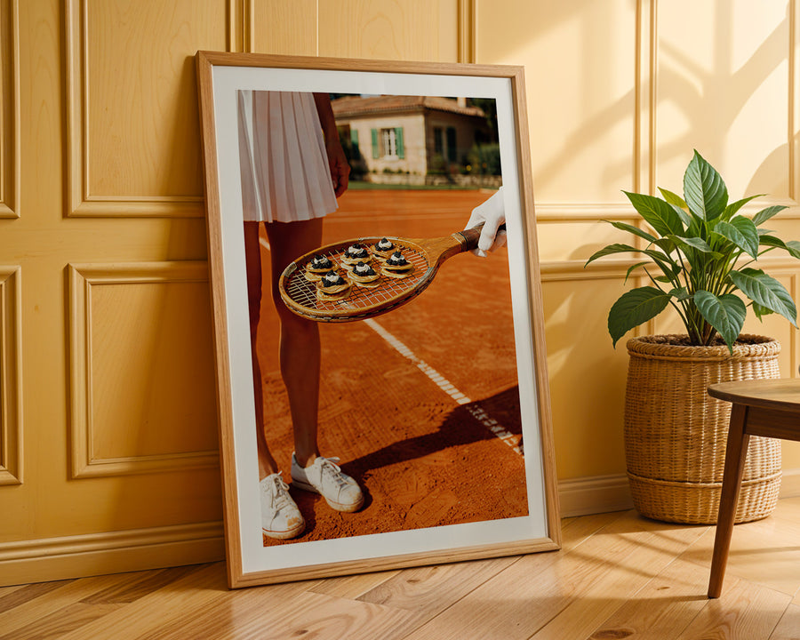 Framed artwork of a person holding a tennis racket with small cakes on a tennis court, leaning against a wooden wall.