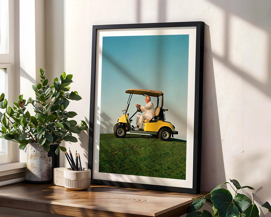 Framed picture of a person in a golf cart on grass, placed on a wooden surface with plants.