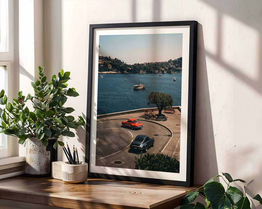 Framed photograph of a scenic road with cars by a body of water, placed on a wooden surface with plants.