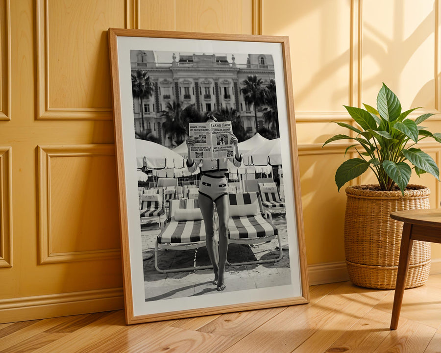 Framed black and white photo of a person on lounge chairs against a building, with a plant and table in the background.