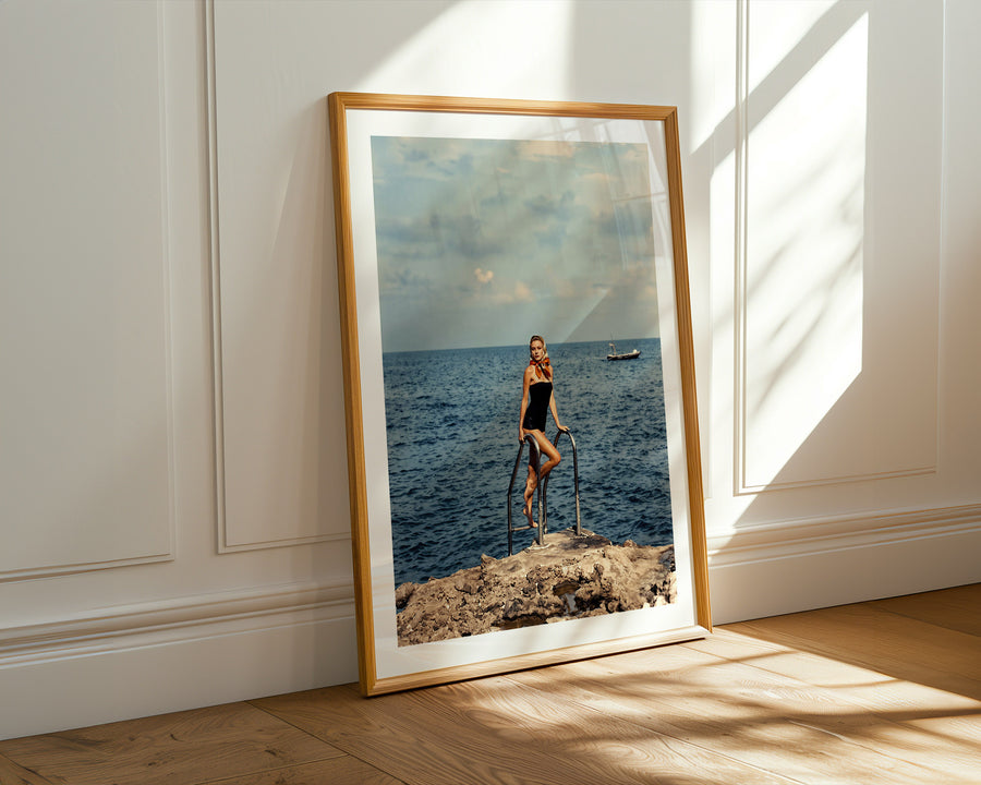 A framed vertical art print leaning against a white paneled wall on a light wood floor, showing a woman standing on a tricycle by the ocean under warm sunlight.