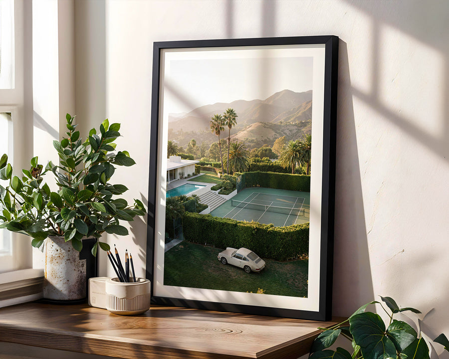 Framed photograph of a tennis court with mountains in the background, placed on a wooden surface with plants.