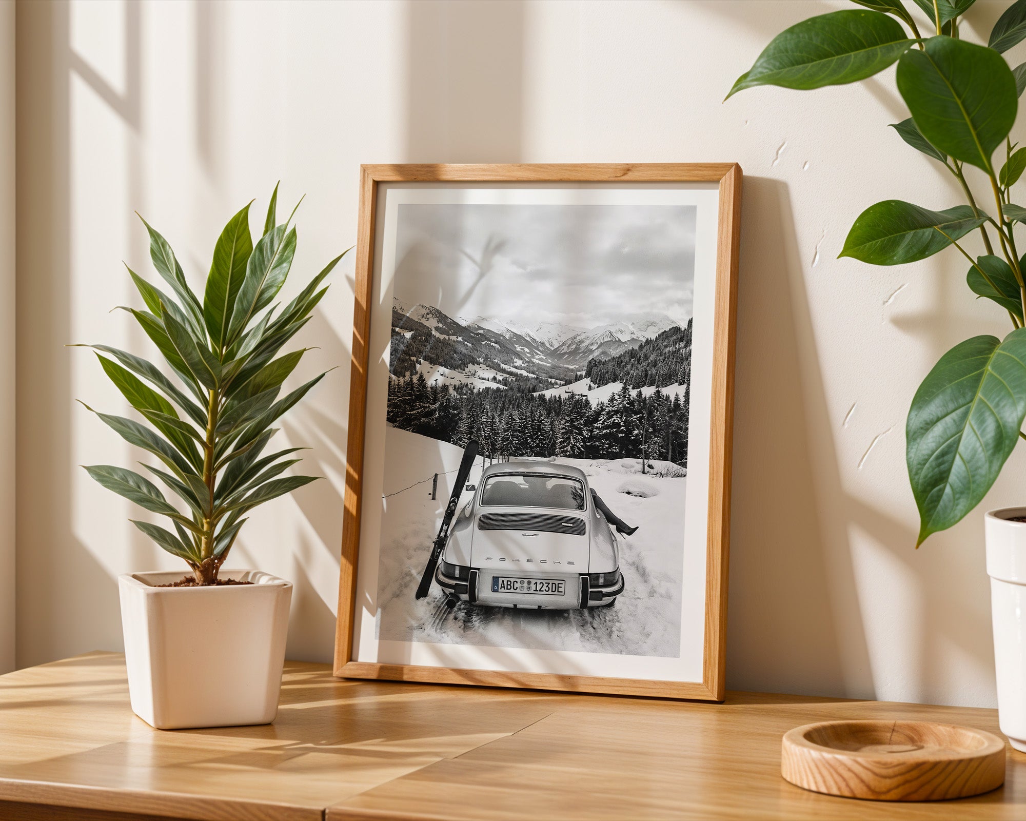 Framed black and white photo of a snowy mountain landscape with a 911 Porsche car, placed on a wooden surface with plants.