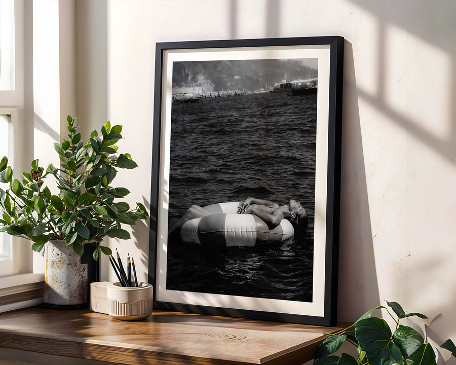Framed black and white artwork of a person on an inner tube in water, placed on a wooden shelf with plants.