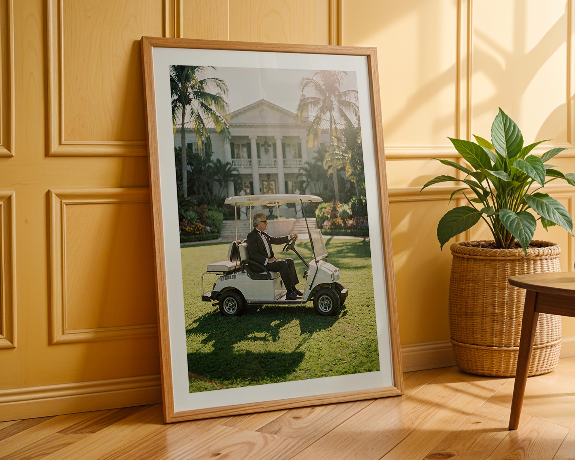 Framed photograph of a person in a golf cart on grass, leaning against a wooden wall with a plant beside it.