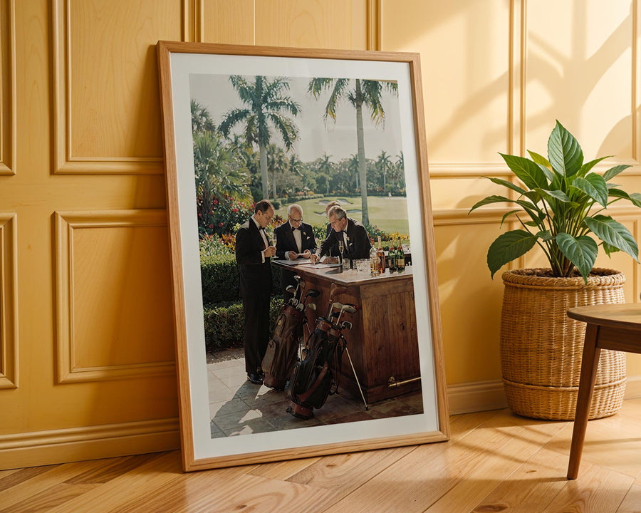 Framed photograph of a group of people at a table outdoors, leaning against a wooden wall with a plant in the foreground.