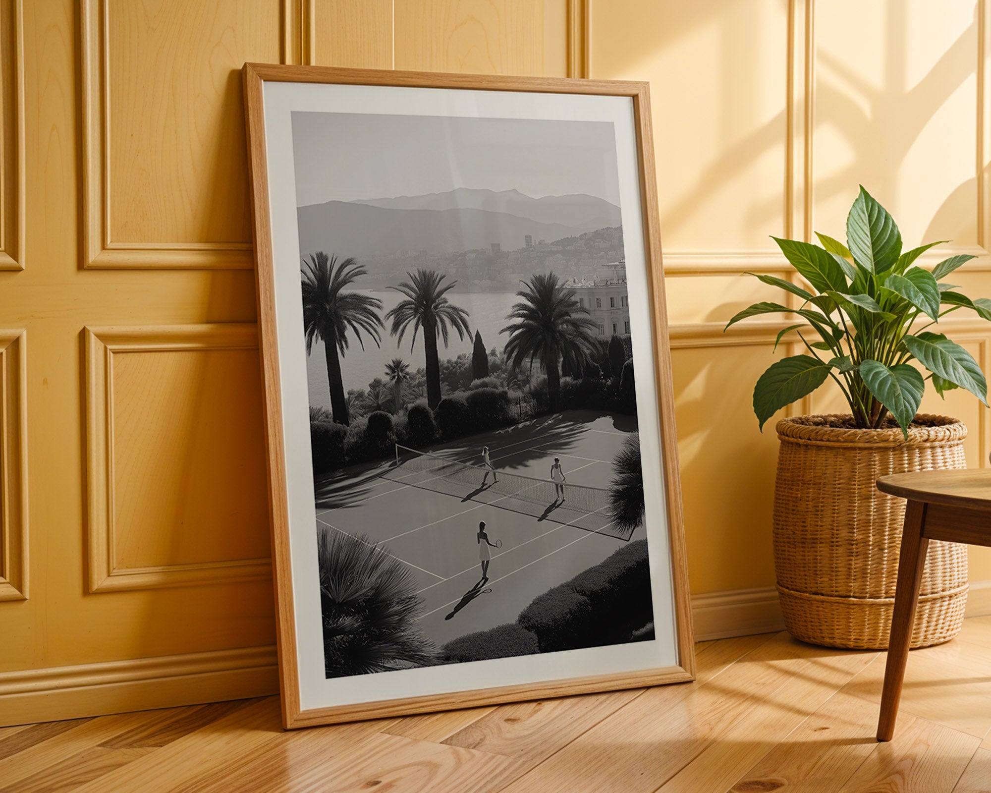 Framed black and white print of a tennis court scene on a wooden floor with a plant in the background.