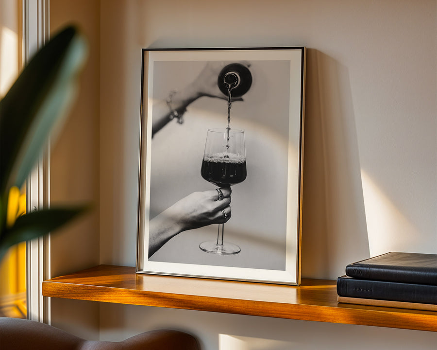 Framed artwork of a hand holding a glass with a black liquid on a wooden shelf.