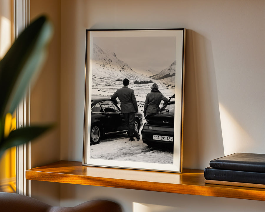Framed black and white photograph of two people by a car on a mountain road, displayed on a wooden shelf.