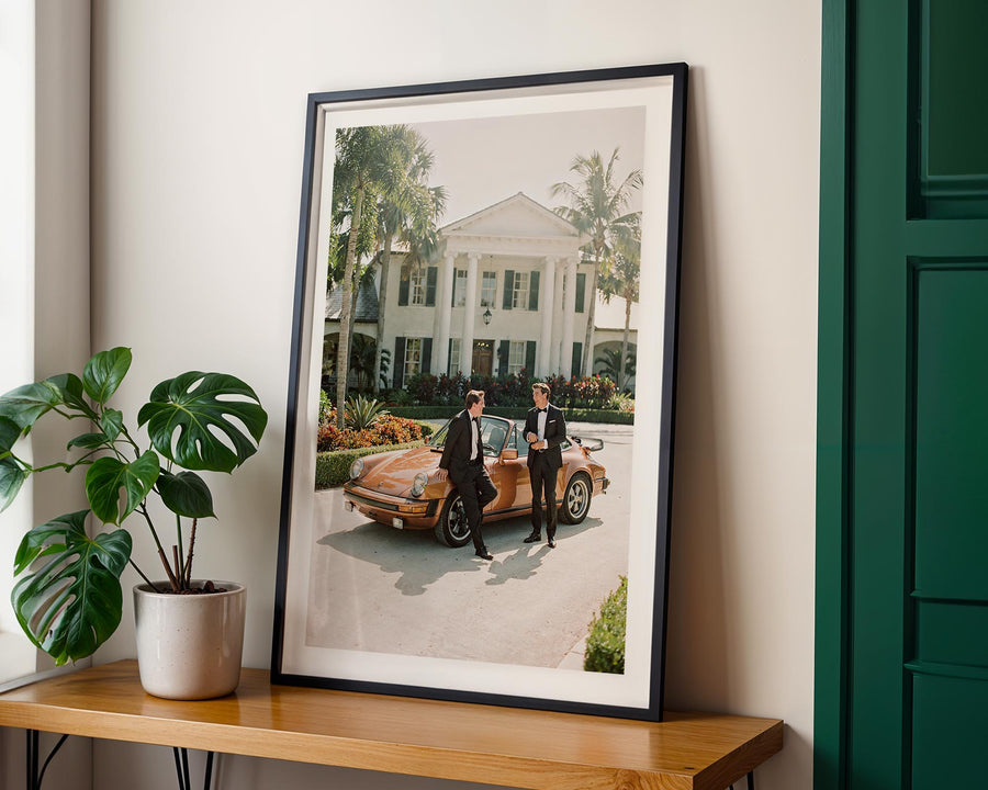 Framed photograph of two people by a vintage car on a wall next to a plant and wooden shelf.