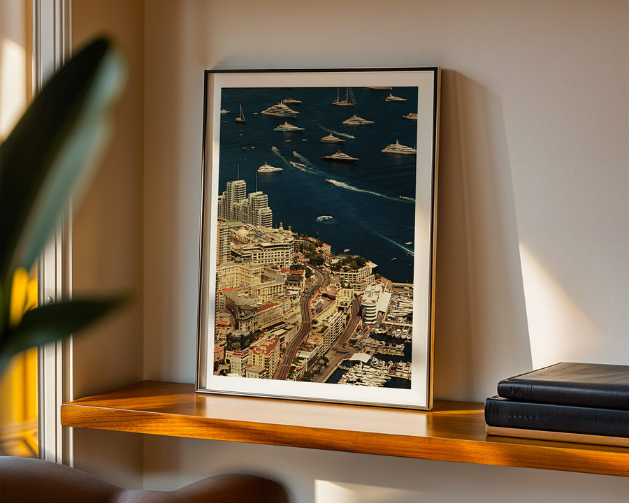 Framed cityscape artwork on a wooden shelf with a plant and books in the background.