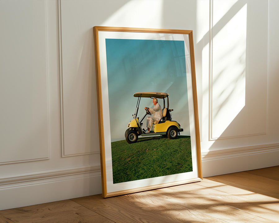 Framed photograph of a person in a golf cart on grass, leaning against a white wall with sunlight casting shadows.