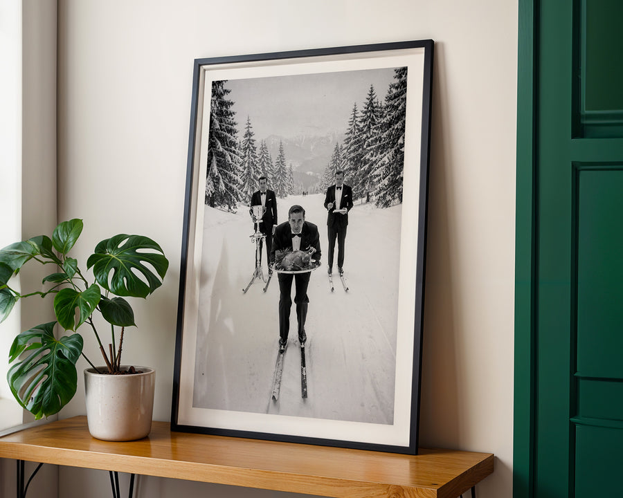Framed black and white photograph of people skiing on a wall above a wooden shelf with a plant.