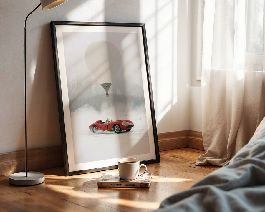 Framed artwork of a red car and green balloon on a wall next to a bed with a lamp and cup.