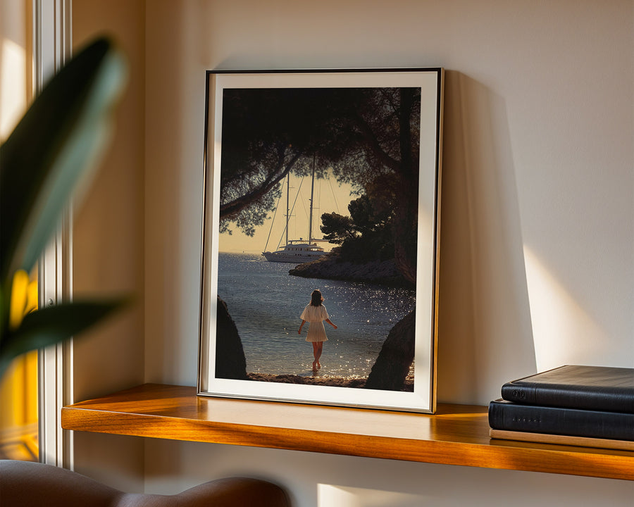 Framed artwork of a woman walking on a beach with sailboats, displayed on a wooden shelf.