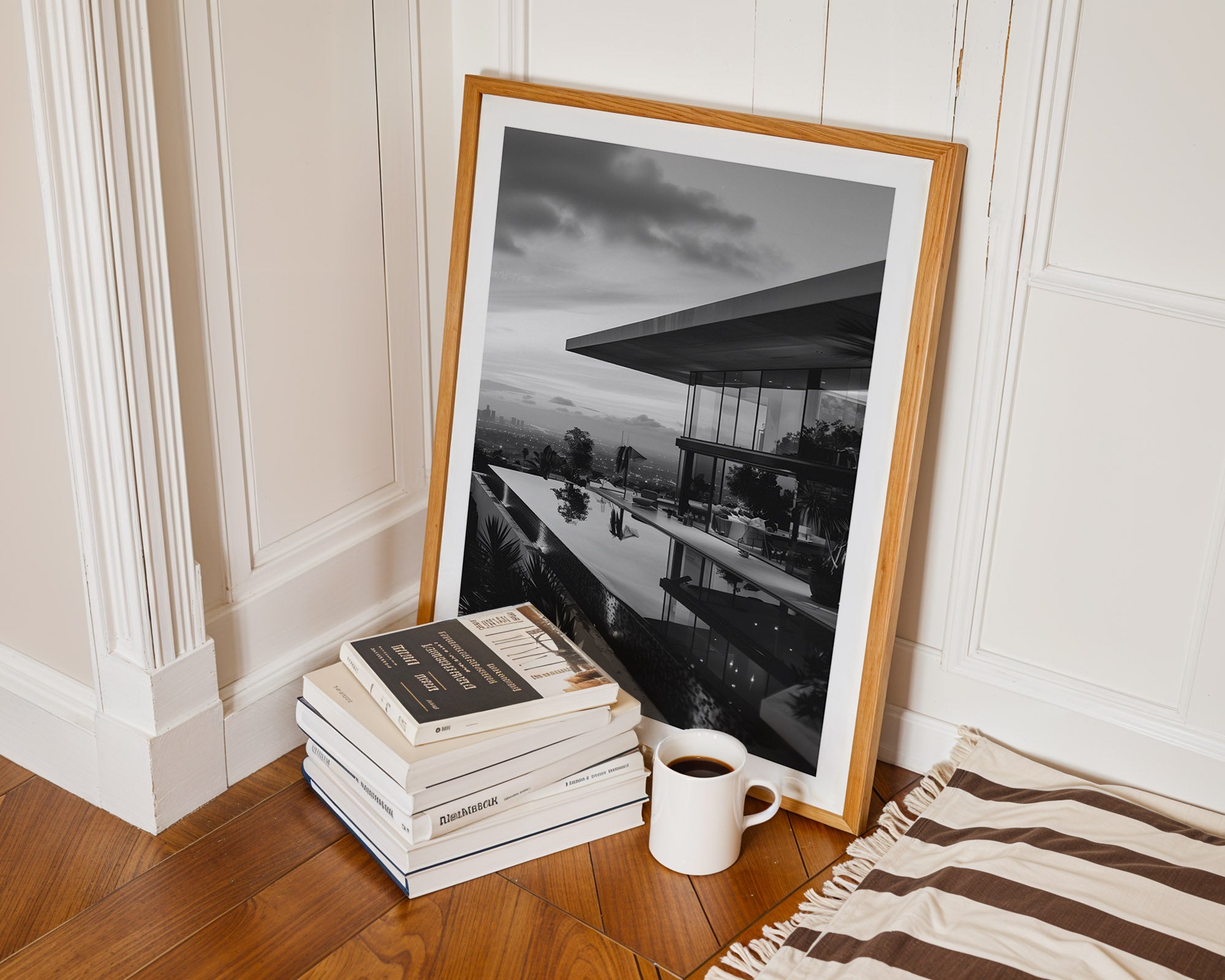 Framed black and white artwork leaning against a wall next to books and a coffee cup on a wooden floor.