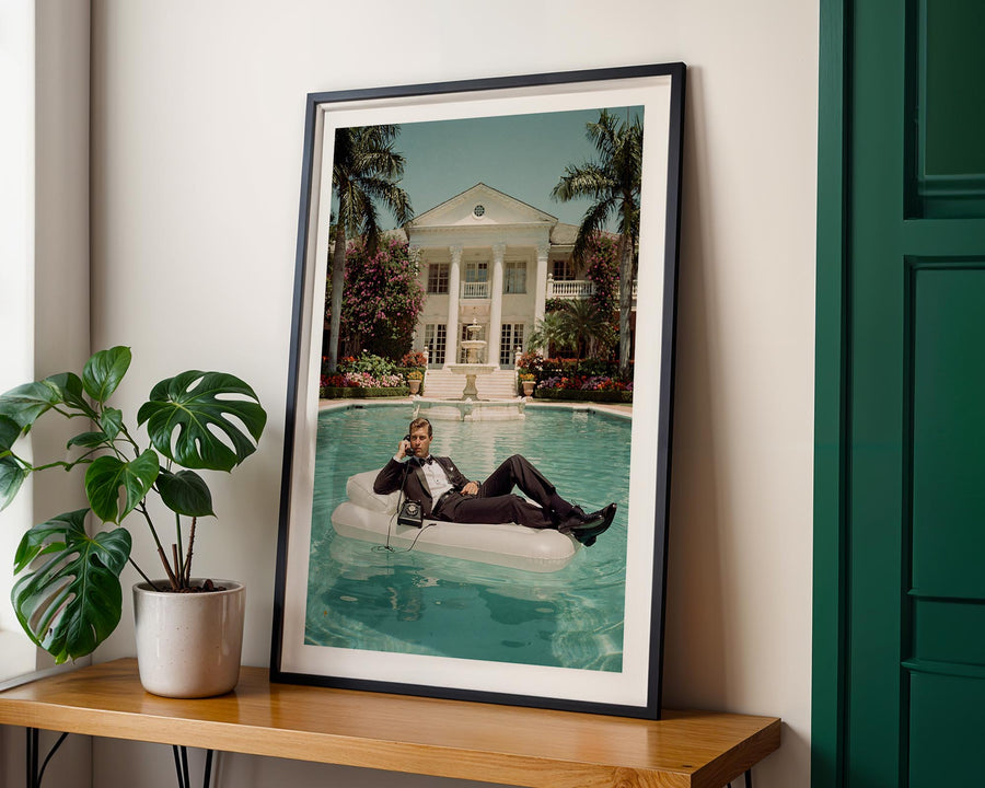 Framed photograph of a person on an inflatable pool float by a large house with palm trees.