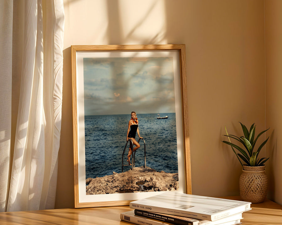 A framed art print depicting a woman standing on a rocky cliff overlooking a blue ocean under a cloudy sky, styled on a wooden table next to a small potted plant with warm sunlight filtering through a curtain.