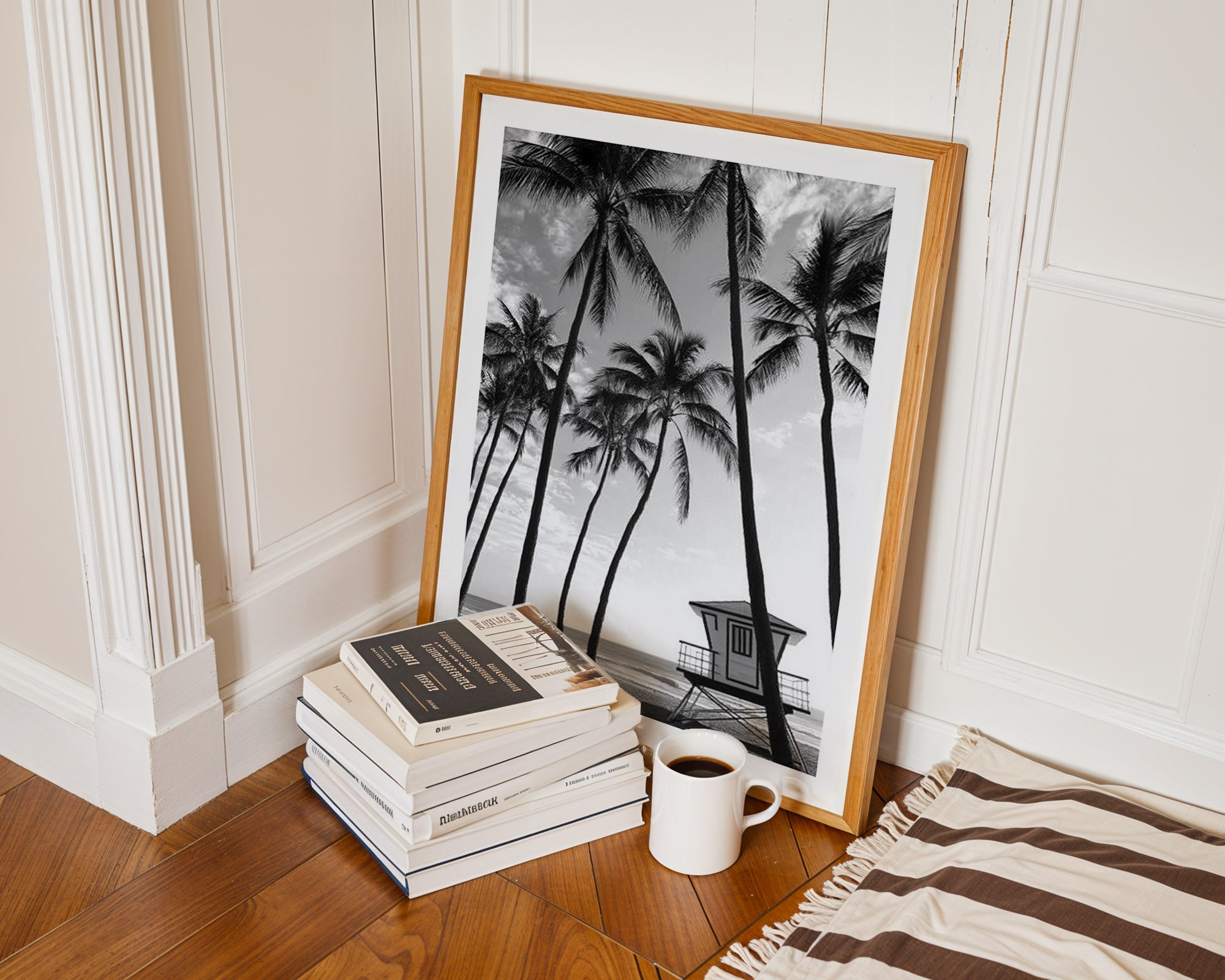 Framed black and white palm tree print on a wooden floor with books and a mug.