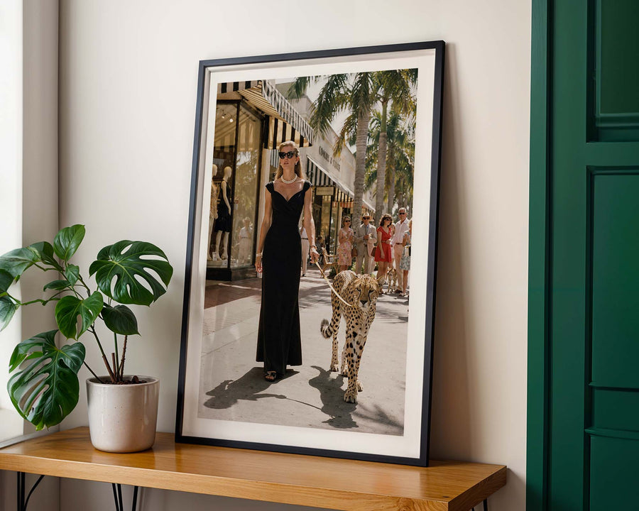 Framed photograph of a woman walking a leopard on a city street, placed on a wooden table with a plant beside it.