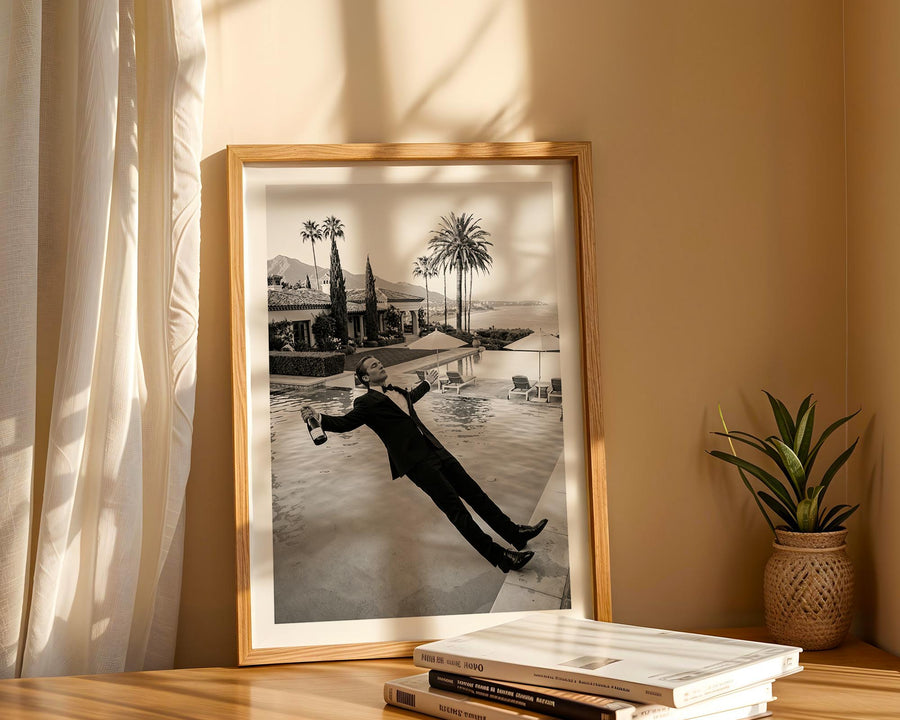 Framed black and white photo of a person by a pool with palm trees, placed on a wooden surface with books and a plant.