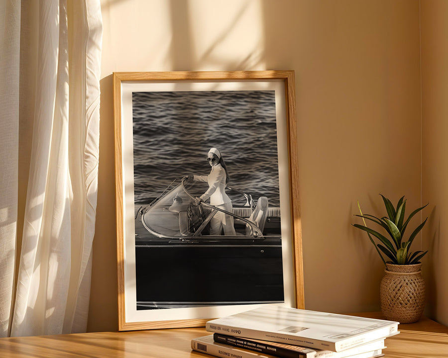 A framed black and white art print depicting a person rowing a boat on water, styled on a wooden table next to a potted plant with warm sunlight filtering through a curtain.
