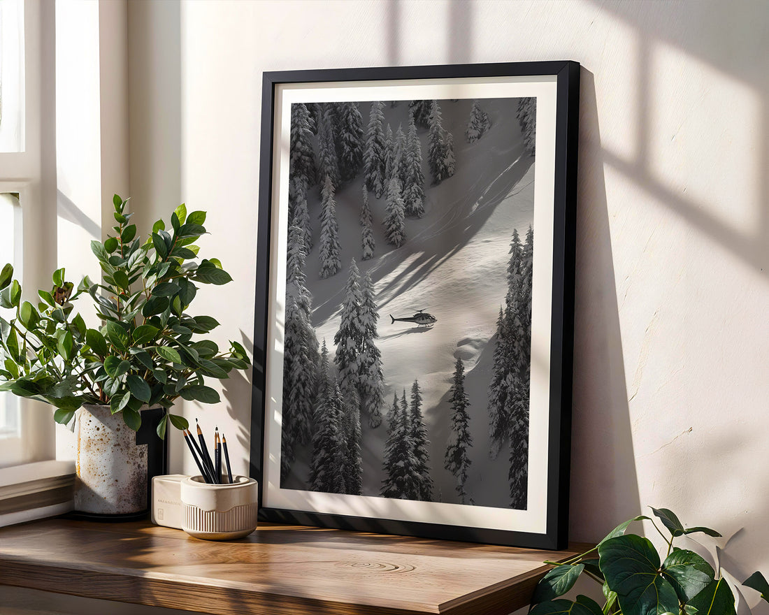 Framed black and white landscape print of a snowy forest with a helicopter, placed on a wooden surface with plants.