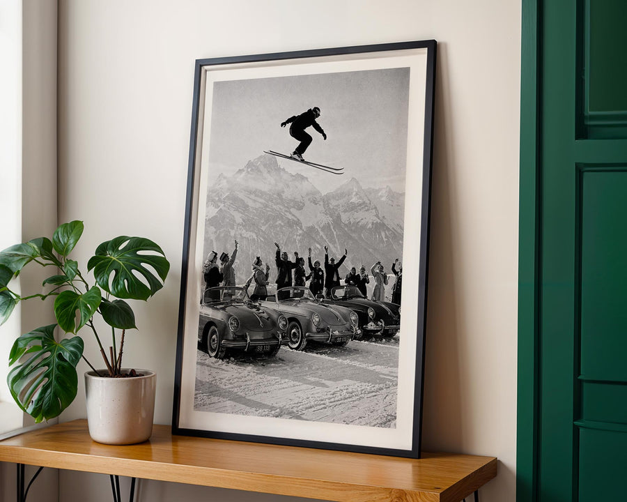 Framed black and white print of a skier in mid-air over a crowd with cars below, on a wall above a wooden shelf with a plant.