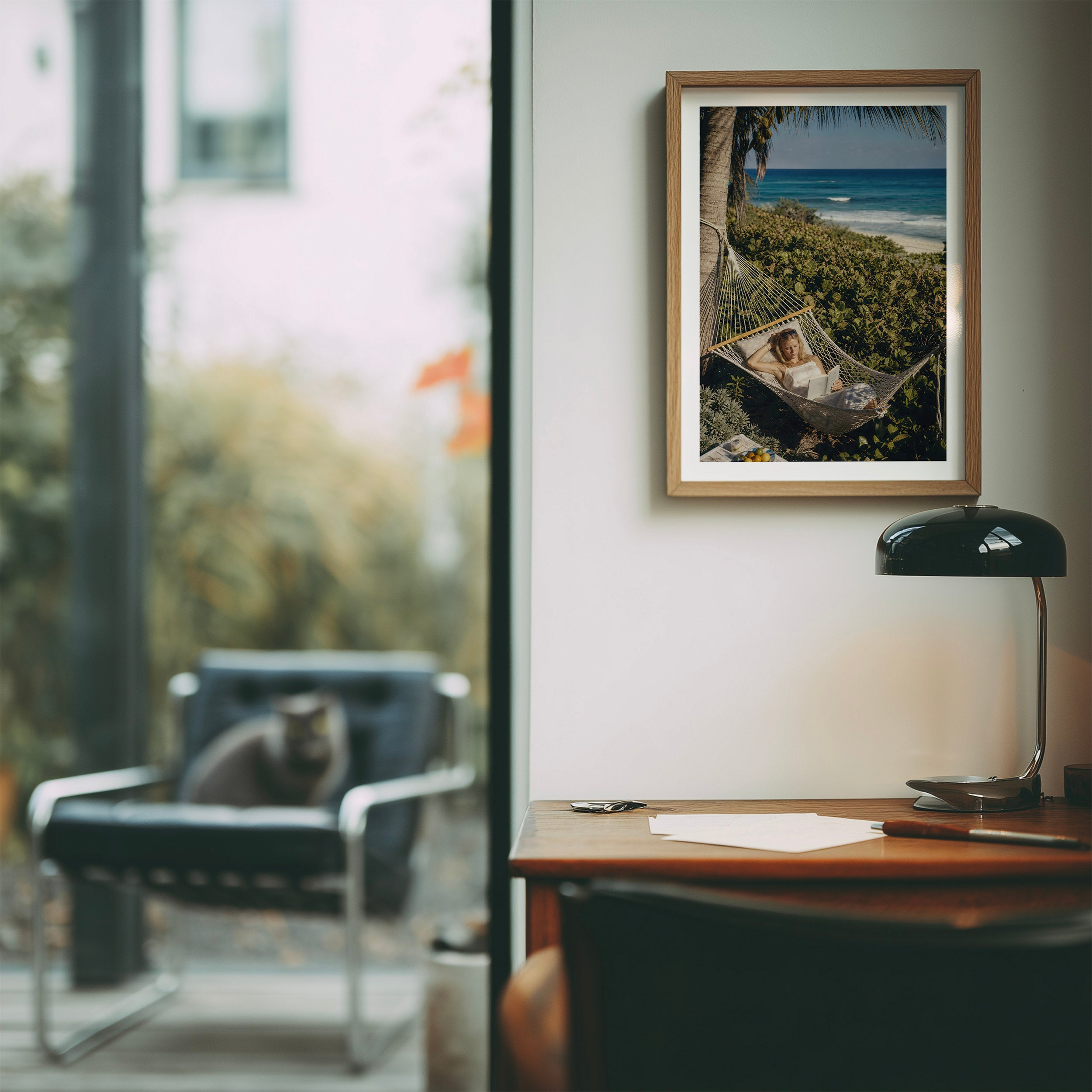 A framed art print depicting a woman relaxing in a hammock by the beach hangs on a light gray wall in a modern office setting. The scene includes a wooden desk with a black lamp, a blurred chair with a cat, and a large window.